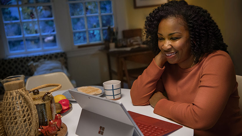 A woman sits at her kitchen table and smiles while looking at a laptop computer.